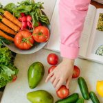 Top view of fresh vegetables and hands arranging them with a recipe book, promoting healthy eating.